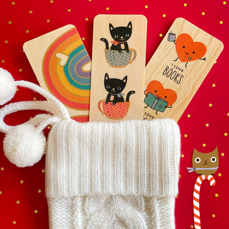 Three birch wood bookmarks in a white sweater stocking on red holiday background. Bookmarks feature a rainbow with small birch heart, black cats wearing scarfs sitting in coffee cups and smiling happy hearts holding books.