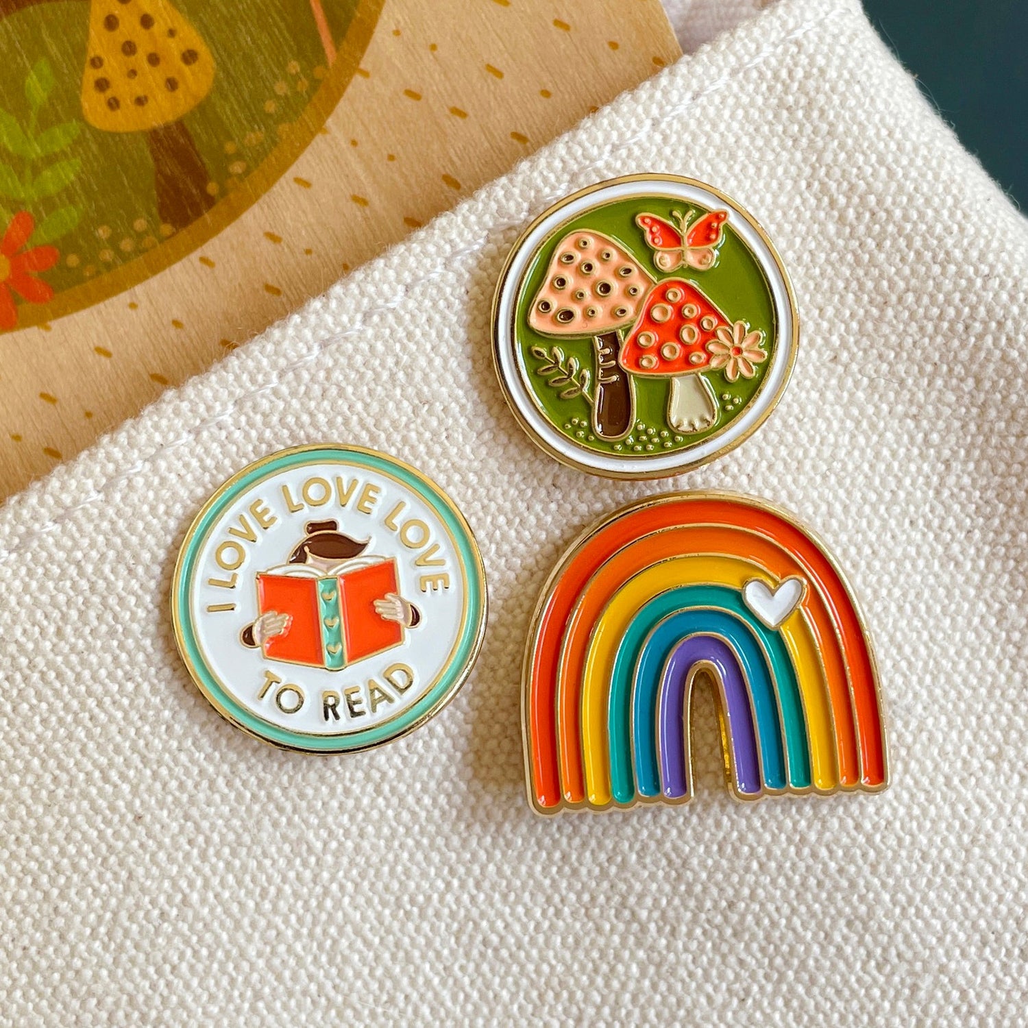 Three enamel pins on a canvas tote. Love to Read is a round pin with a head behind a book. Another pin is a folksy pin and red mushrooms with olive green background and pink butterfly. And a colorful rainbow shaped pin with a small white heart.