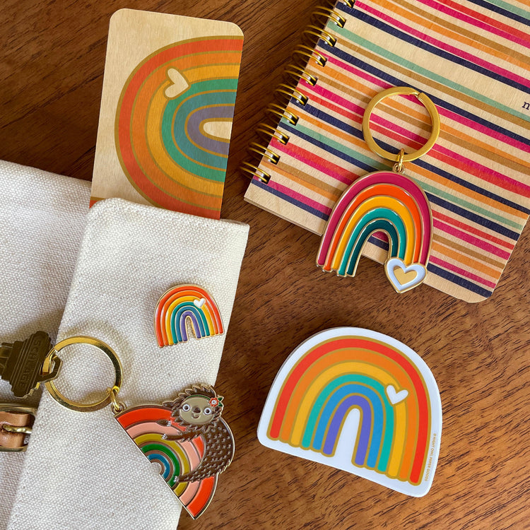 Rainbow Sloth and Rainbow keychains alongside a Rainbow Vibes enamel pin and vinyl sticker