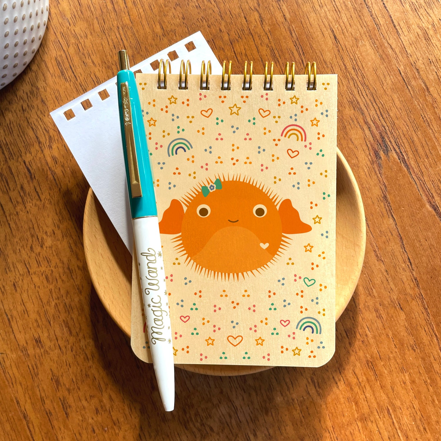 Wood mini notepad with cute orange puffer fish displayed with pen with gold details that reads 'Magic Wand'. Background pattern on notepad has a fun retro vide with mini hearts and rainbows.