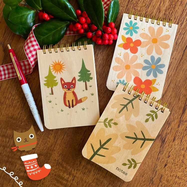 Three small notepads with birch wood cover featuring nature designs with cute fox sitting in the woods, big, colorful modern floral pattern and bold neutral tan and green pattern with leaves.