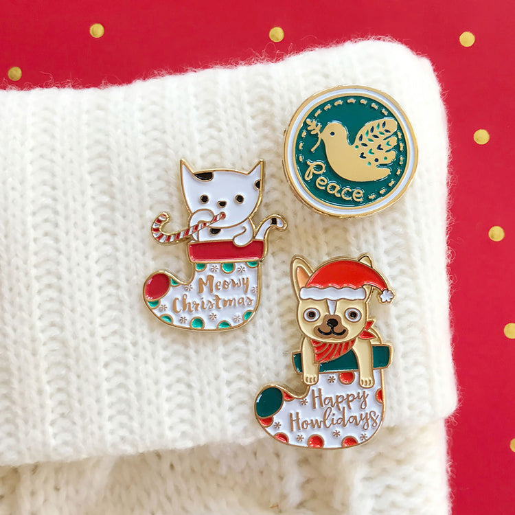 Group of three holiday enamel pins on white stocking. Meowy Christmas with cat in stocking. Happy Howlidays with french bulldog wearting santa hat in stocking. Round pin with blue background featuring a unique modern gold dove and handwritten word Peace.