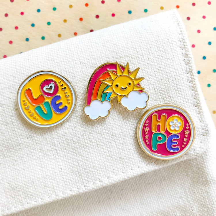 Three bright and colorful enamel pins on a canvas bag. Two are round. One reads Love. One reads Hope. And one is a rainbow with happy sun and puffy clouds.