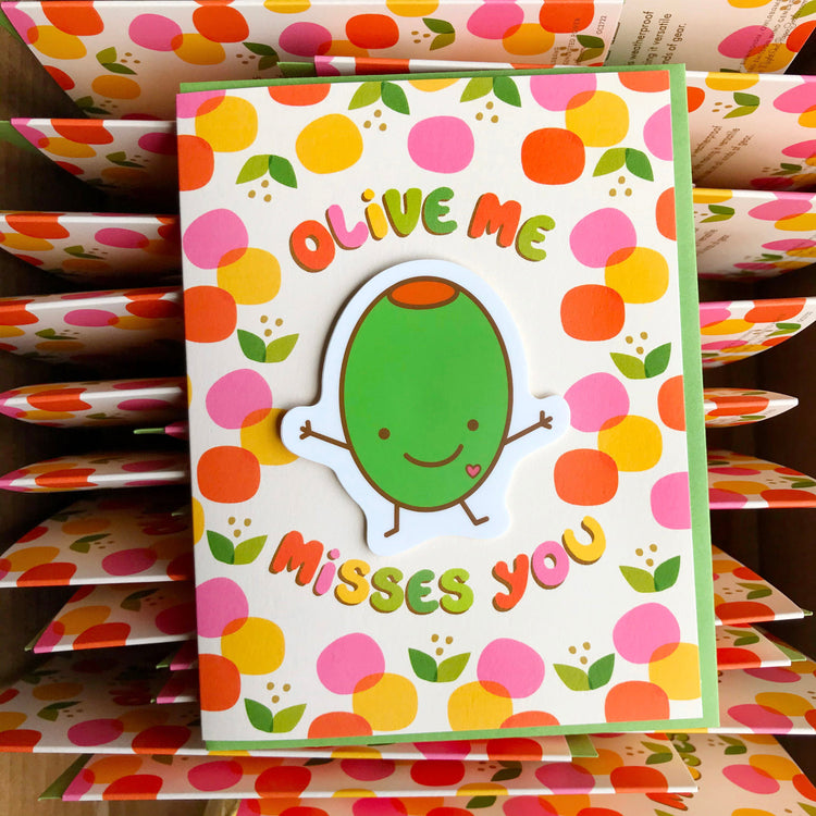Green olive with happy face and stick arms reaching out with a hug. Olive is a removable sticker. Card hasyellow pink, red happy dots. Card reads Olive Me Misses You