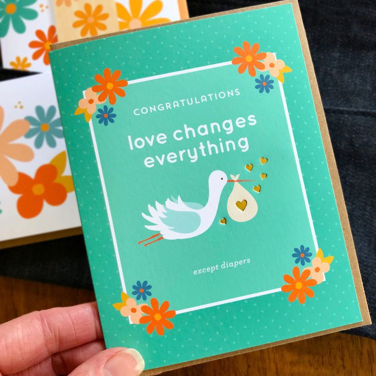 Aqua green congratulations card for new parent. Card has illustration of cute white stork with light blue wings carrying a baby bundle with gold foil hearts and simple flowers suitable for either a boy or girl.
