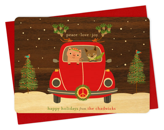 Customizable birch wood holiday card with fun illustration of a pig and fox riding in a red car with peace symbol and wreath on the front of the car with a printed walnut wood grain sky. Card features “Peace Love Joy” sentiment and a personal greeting. Shown with a red envelope.