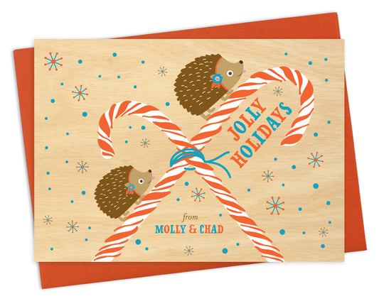 Customizable paper holiday card featuring hedgehogs and candy canes on a snowy printed birch wood grain background with "Jolly Holidays" text and a personal greeting. Shown with a red envelope.