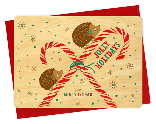 Customizable wood holiday card featuring hedgehogs and candy canes on a snowy birch wood grain background with "Jolly Holidays" text and a personal greeting. Shown with a red envelope.
