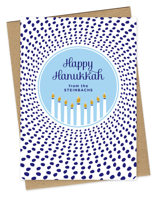 Customizable paper Hanukkah card featuring a glowing menorah inside a light blue medallion that reads “Happy Hanukkah” along with a personal greeting. The medallion is surrounded by scattered dark blue dots. Shown with a kraft envelope.