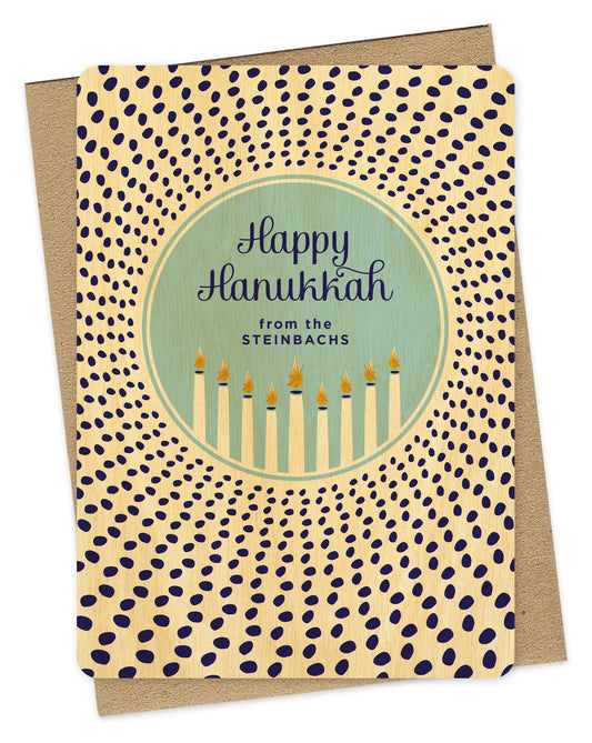 Customizable wood Hanukkah card featuring a glowing menorah inside a light blue medallion that reads “Happy Hanukkah” along with a personal greeting. The medallion is surrounded by scattered dark blue dots. Shown with a kraft envelope.