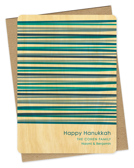 Customizable wood Hanukkah card featuring vibrant blue interwoven stripes, “Happy Hanukkah” text and a personal greeting on a birch wood background. Shown with a kraft envelope.