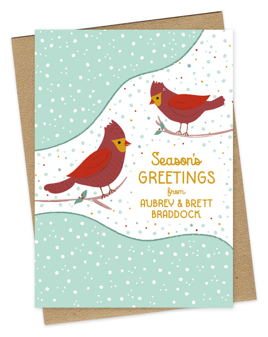 Customizable paper Christmas card featuring two red cardinals perched against a snowy background with “Season’s Greetings” text and a personal greeting. Shown with a kraft envelope.