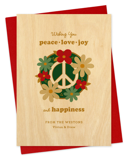 Customizable paper holiday card featuring a bold peace sign decorated with festive florals, a “Wishing you peace, love, joy and happiness” text and a personal greeting. Background is printed birch wood grain. Shown with a red envelope.