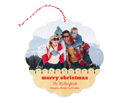 Front of customizable paper holiday ornament card featuring a photo with a festive Christmas lights border and “Merry Christmas” text and a personal message on a printed birch wood background. Round ornament with scalloped edges.