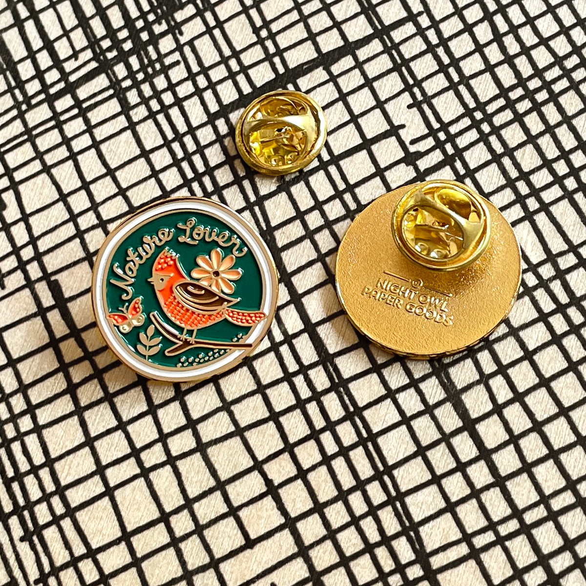 A round enamel pin shown from both front and back on a black-and-white crosshatch background. The front features a blue background with a red and brown bird, floral design and 'Nature Lover' text, outlined in gold. The back is all gold with a butterfly clasp and Night Owl Paper Goods logo.