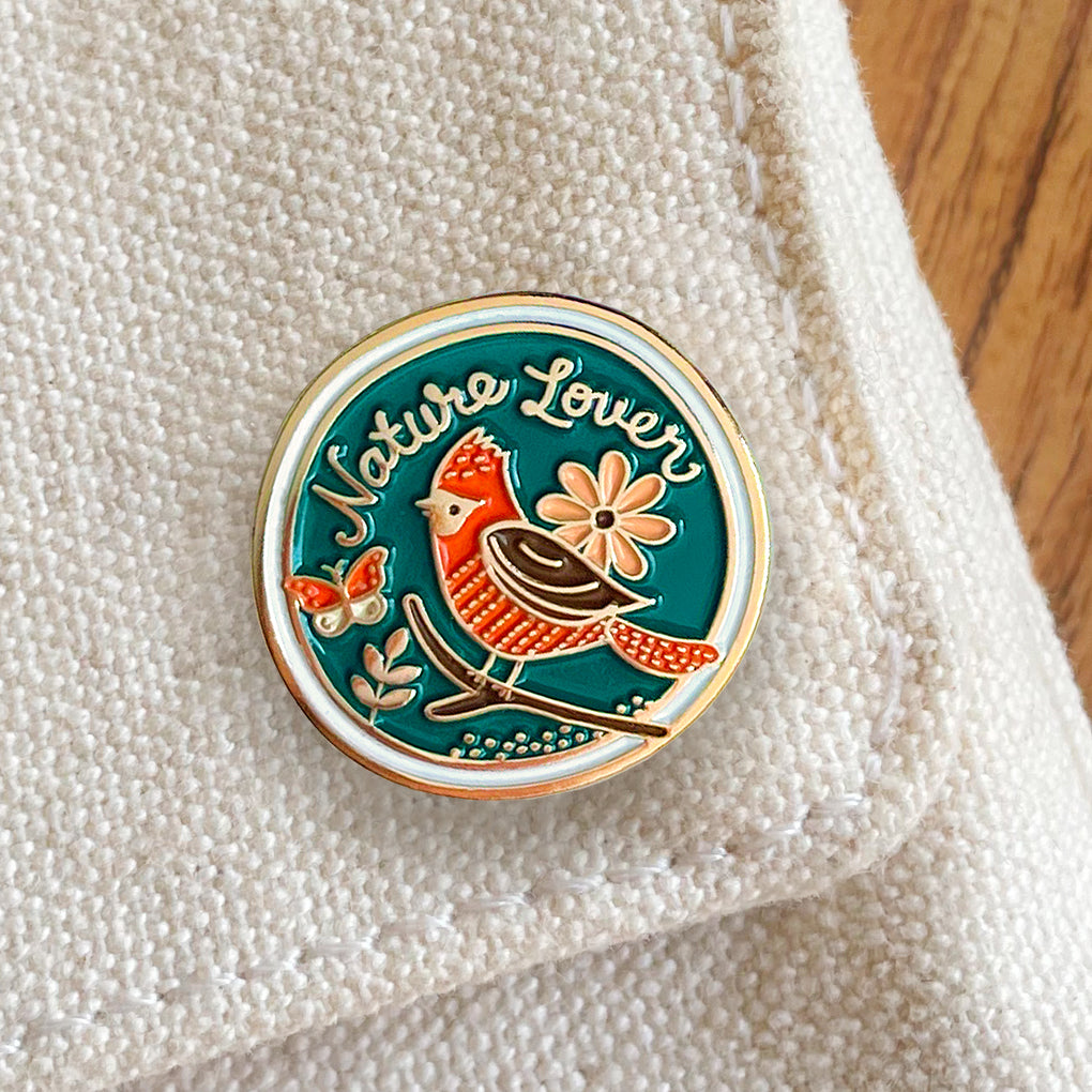 A round enamel pin with a gold border and a blue background, featuring a red and brown bird, a peach flower, a butterfly and the words 'Nature Lover' in gold script. The pin is placed on a textured cream fabric next to a wooden surface.