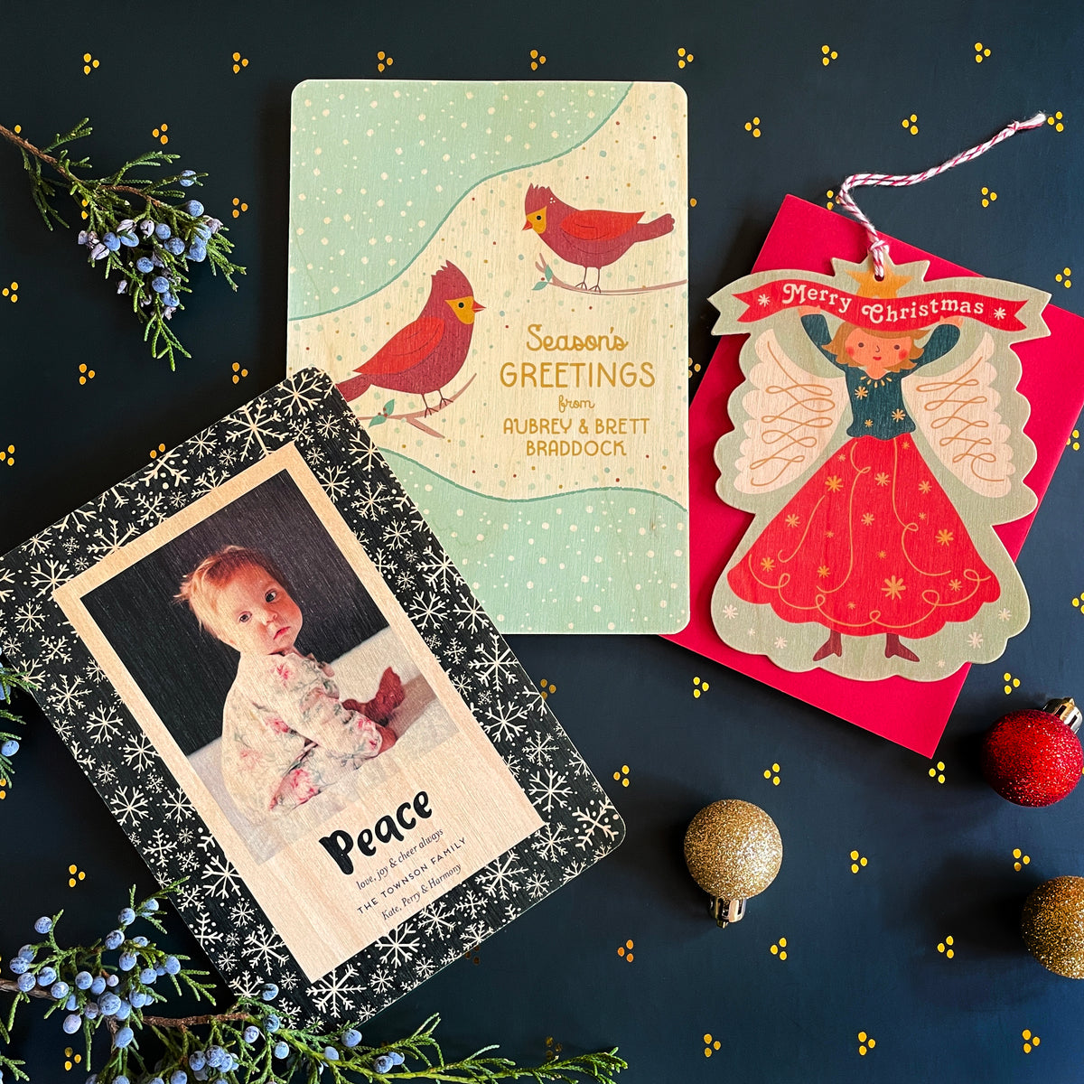 Three customizable birch wood holiday cards. 'Peace' card with photo and personal sentiment with black background and illustrated snow flakes. 'Season's Greetings' card with two red cardinals and snowy dot pattern with custom text on front and an ornament in the shape of an angel with 'Merry Christmas' greeting and customizable back.