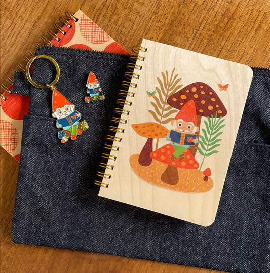 Grouping of Reading Gnome items. A key chain, an enamel pin and a wood notebook with happy gnome sitting on a mushroom while reading a book in a forest setting.