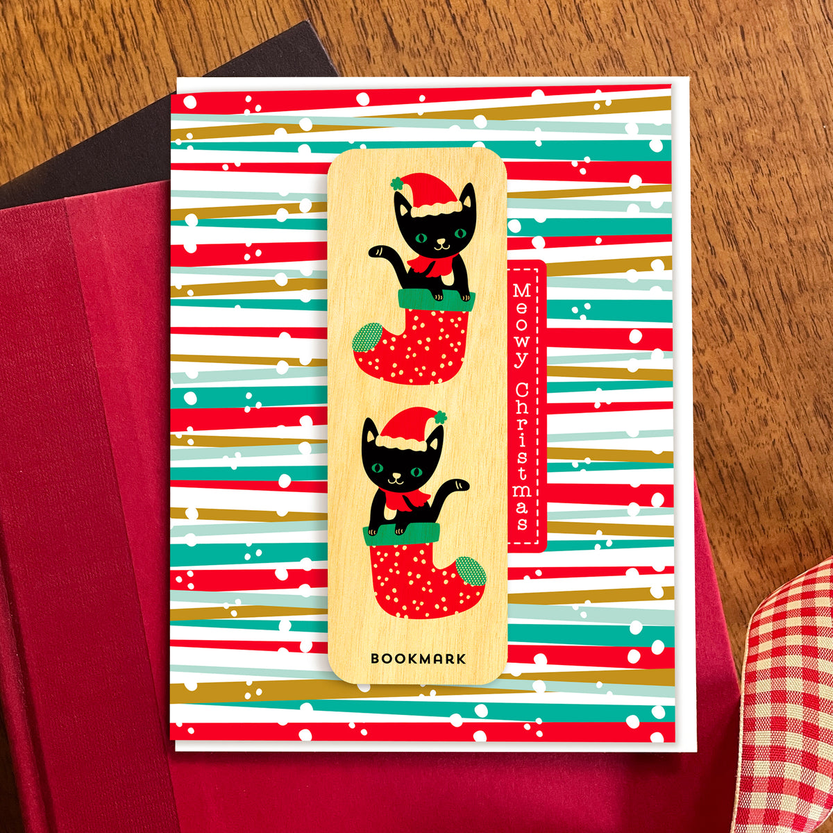 Folded Christmas greeting card with colorful red, green and gold striped background, “Meowy Christmas” text and a removable birch wood bookmark featuring two black cats in red Christmas stockings. Pictured with a white envelope  and sitting atop a book on a wooden table with a red gingham ribbon.