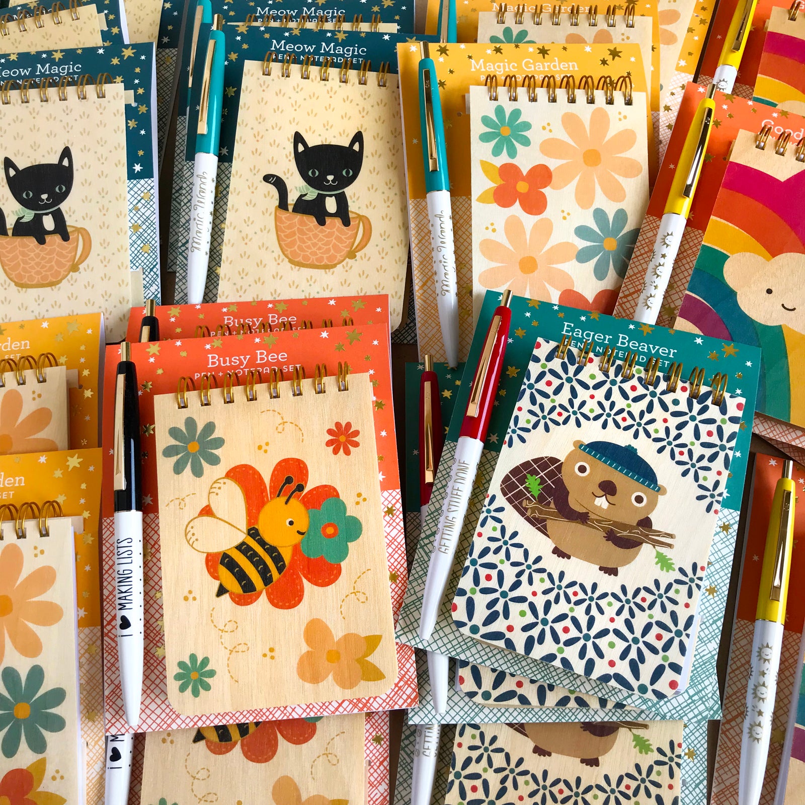 Collection of small wood notebooks and pens with fun sayings features black cat in coffee cup with magic wand pen, modern floral design, rainbow with happy cloud, busy bee loves making lists,  eager beaver getting stuff done