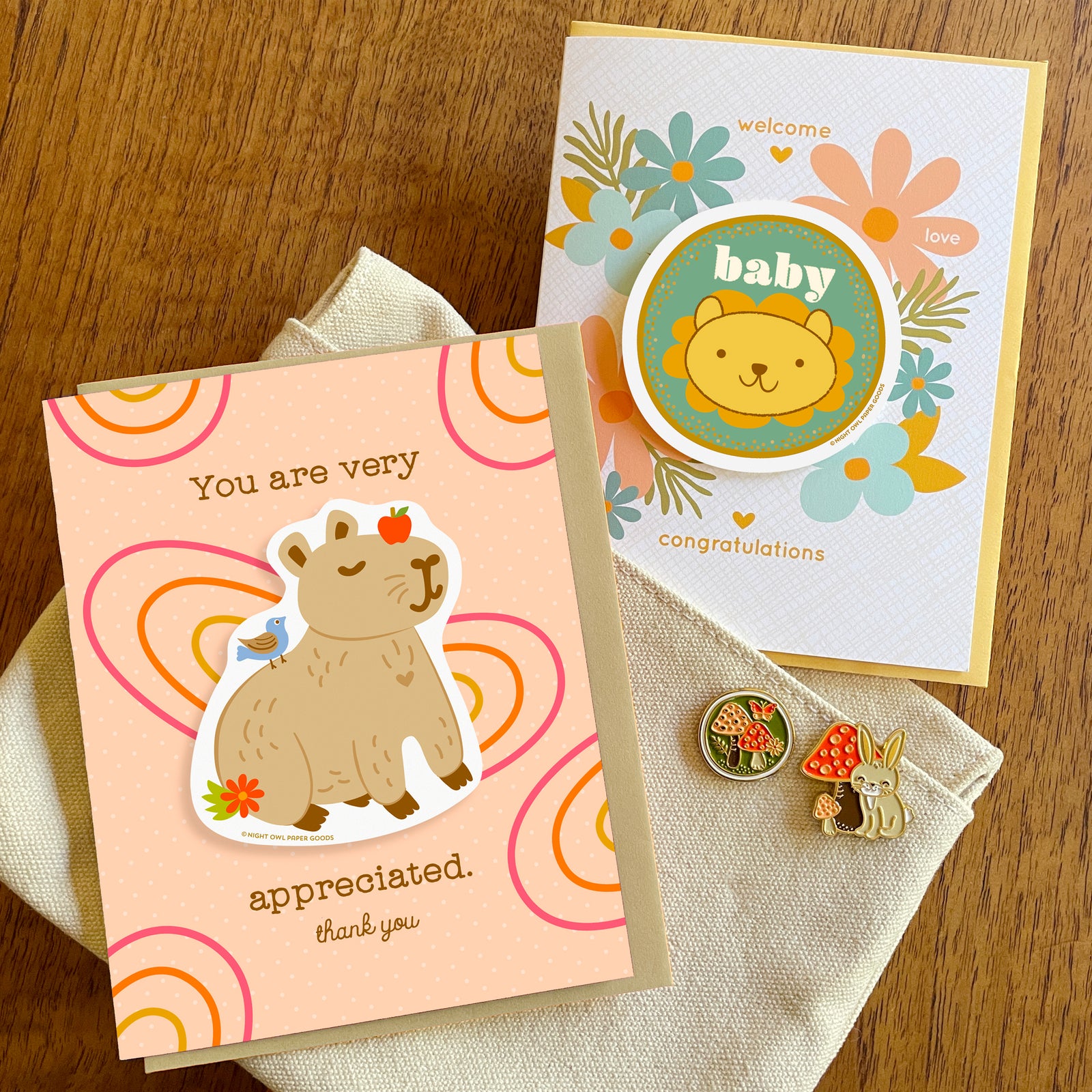 Thank You greeting card with a removable capybara vinyl sticker and the message “You are very appreciated. Thank You”  alongside a baby shower greeting card with a removable baby lion vinyl sticker and "Welcome, Congratulations" text. Also shown are a mushrooms enamel pin and a rabbit enamel pin.
