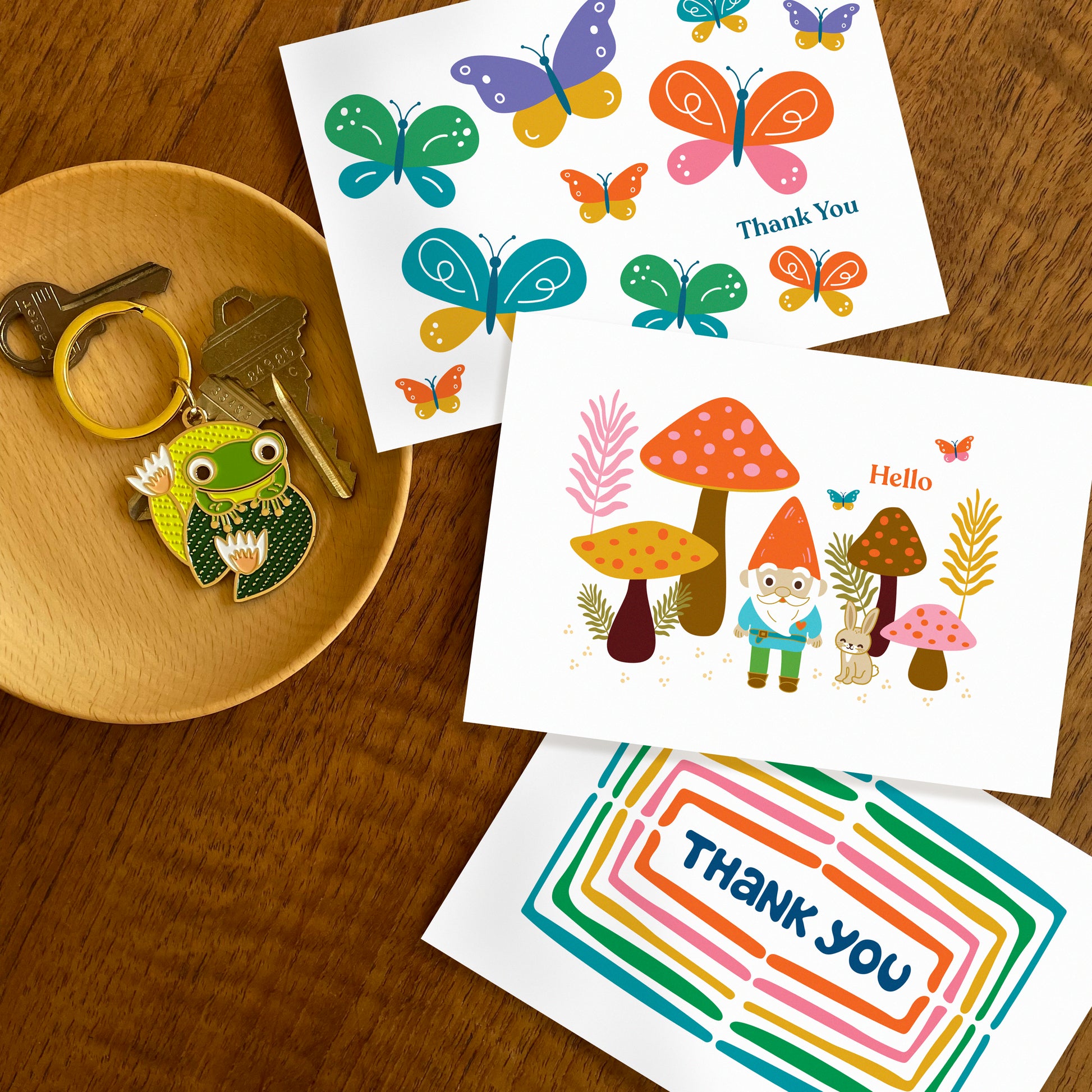 Three folded thank you cards colorful butterflies, gnome in forest with mushrooms and rabbit, and a rainbow of colors surrounding a hand drawn word 'thank you'. Frog on lily pad keychain also featured