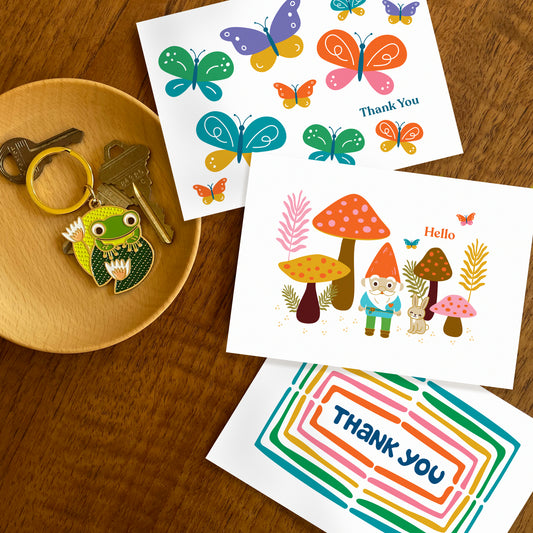 Three folded thank you cards colorful butterflies, gnome in forest with mushrooms and rabbit, and a rainbow of colors surrounding a hand drawn word 'thank you'. Frog on lily pad keychain also featured