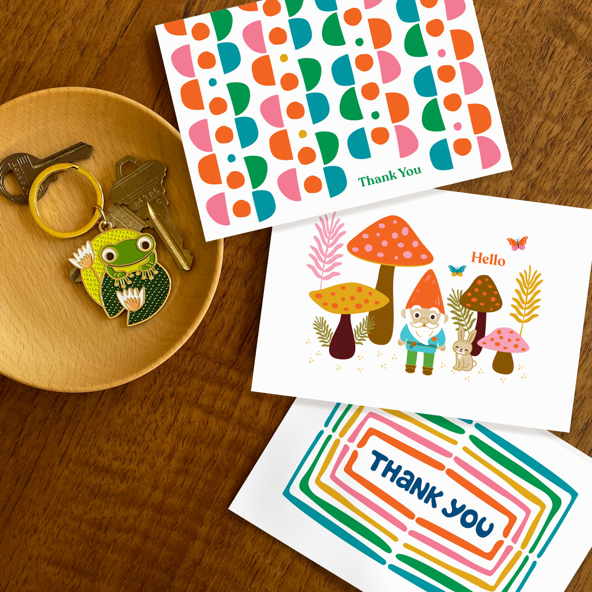Three folded thank you cards irregular hand drawn shapes, gnome in forest with mushrooms and rabbit saying 'hello', and a rainbow of colors surrounding a hand drawn word 'thank you'. Frog on lily pad keychain also featured.