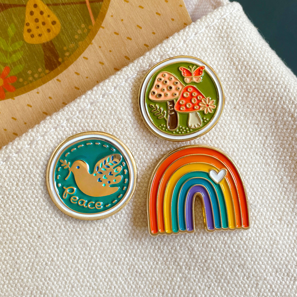 Night Owl Paper Goods three colorful enamel pins: a round teal pin with a gold dove bird and the word 'Peace', a round green pin with red and pink mushrooms and a butterfly, and a rainbow pin with red, orange, yellow, green, blue, and purple arcs and a small white heart.