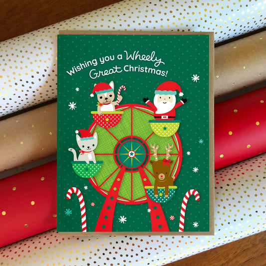 Christmas card with Santa, reindeer, dog and cat on a Ferris wheel with 'Wishing you a Wheely Great Christmas!' greeting. Card sits atop festive wrapping paper.