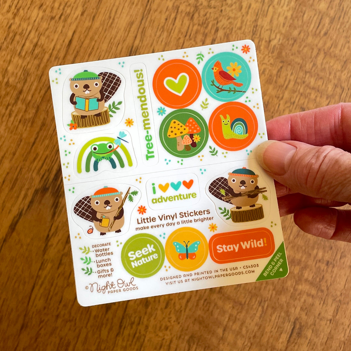 Hand holding woodland animal kiss-cut vinyl stickers featuring beavers, a frog, mushrooms, a bird, a snail and playful sayings in warm earthy colors. Made for decorating water bottles, lunch boxes, gifts and more. Designed and printed in the USA.