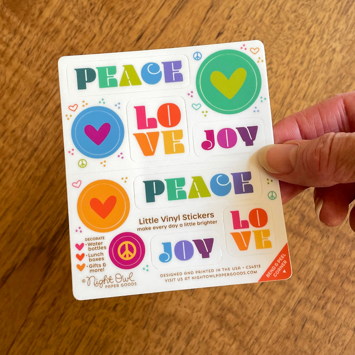 Hand holding a sheet of colorful kiss-cut vinyl stickers on a white background. The stickers feature hearts, peace signs and “PEACE LOVE JOY” designs in various shades of vibrant teal, green, blue, orange, pink, purple and yellow. Text on the sheet reads “Little Vinyl Stickers - make every day a little brighter - Decorate water bottles, lunch boxes, gifts & more - Night Owl Paper Goods - Designed and printed in the USA.”