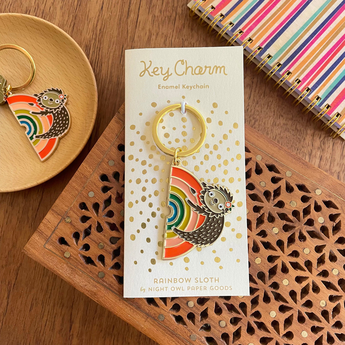 Rainbow Sloth enamel keychain on gold foil stamped light yellow backer card reads "Key Charm Enamel Keychain" at top and "Rainbow Sloth by Night Owl Paper Goods" at bottom.
