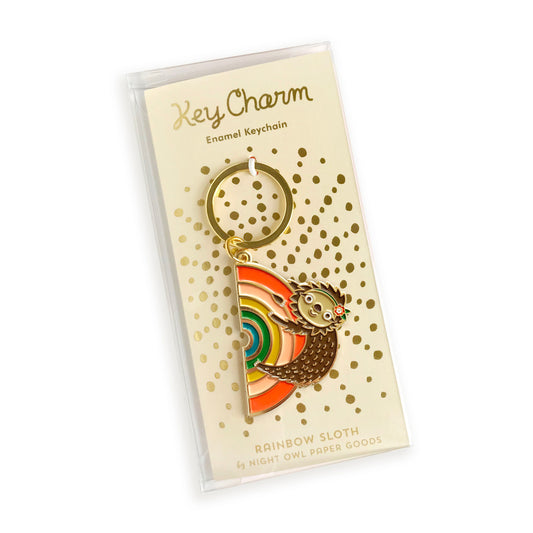 Rainbow Sloth enamel keychain on gold foil stamped backer card packaged in a clear gift box. Light yellow backer card reads "Key Charm Enamel Keychain" at top and "Rainbow Sloth by Night Owl Paper Goods" at bottom.