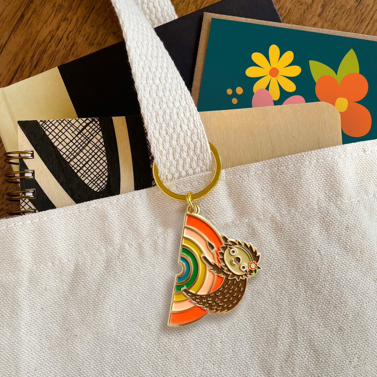 Rainbow Sloth enamel keychain hanging from a canvas tote bag handle