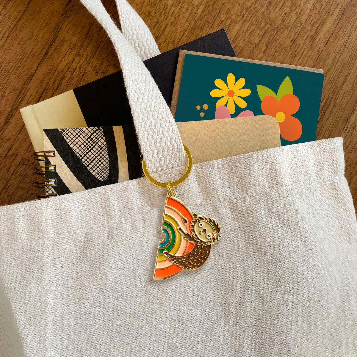 Rainbow Sloth enamel keychain hanging from a canvas tote bag handle