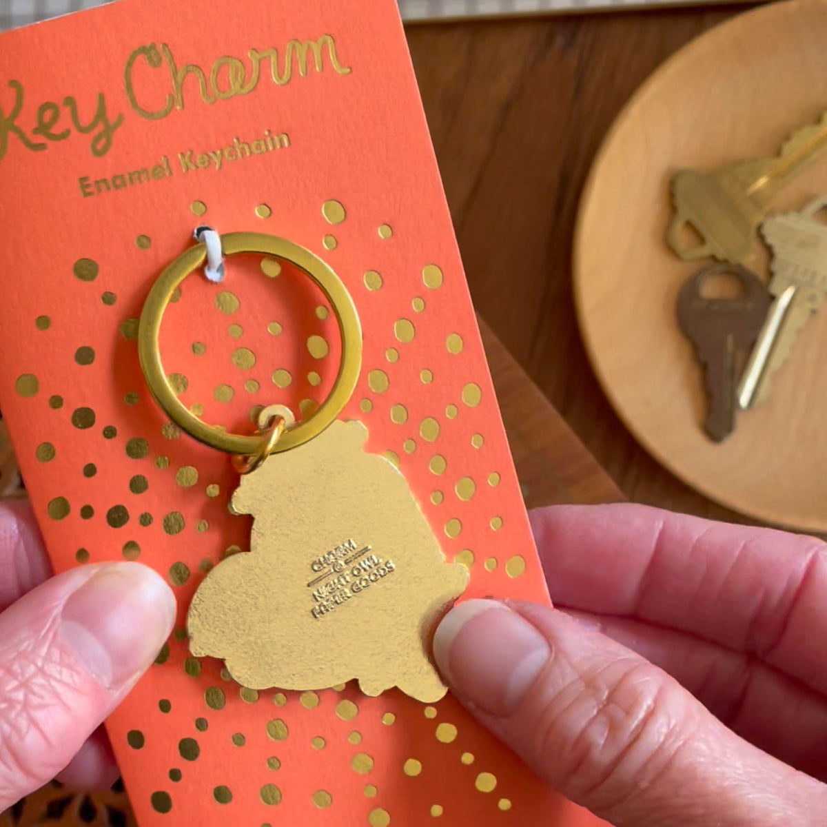 Back of otter enamel keychain on gold foil stamped orange backer card. Backer card reads "Key Charm Enamel Keychain."  Back of keychain is gold and reads Night Owl Paper Goods."