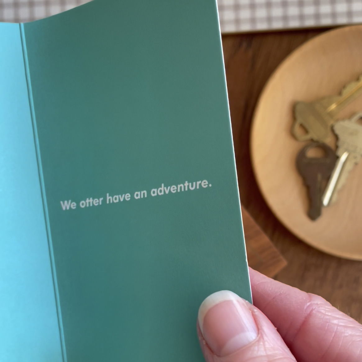 Inside of Otter enamel keychain backer card, which is aqua and reads "We otter have an adventure."