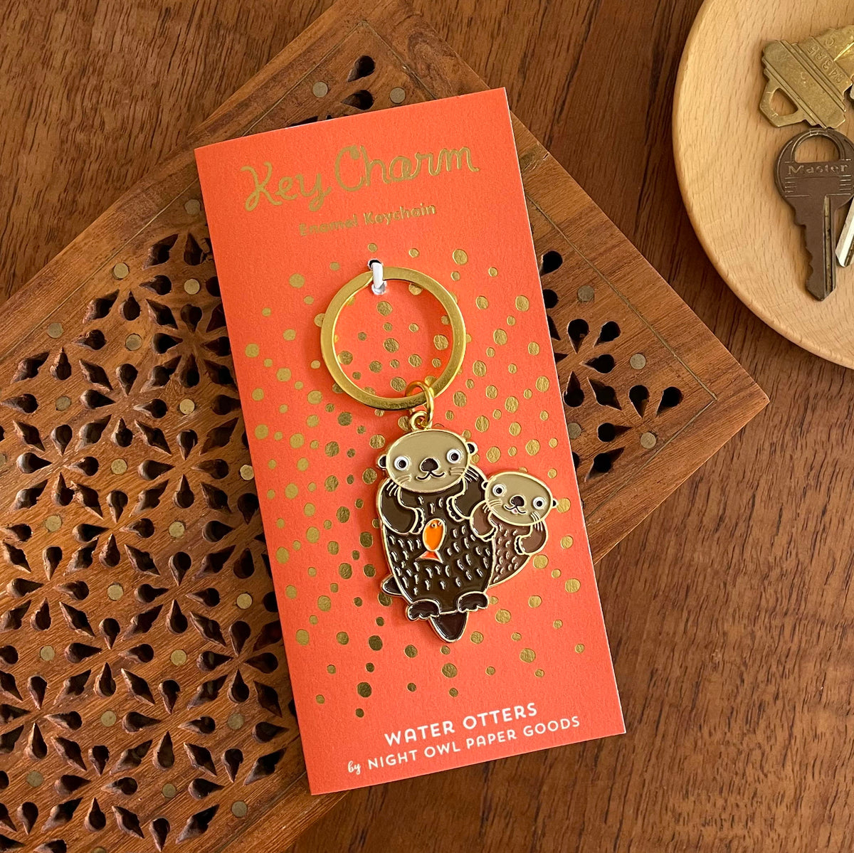 Otter enamel keychain on gold foil stamped orange backer card. Backer card reads "Key Charm Enamel Keychain" at top and "Water Otters by Night Owl Paper Goods" at bottom.