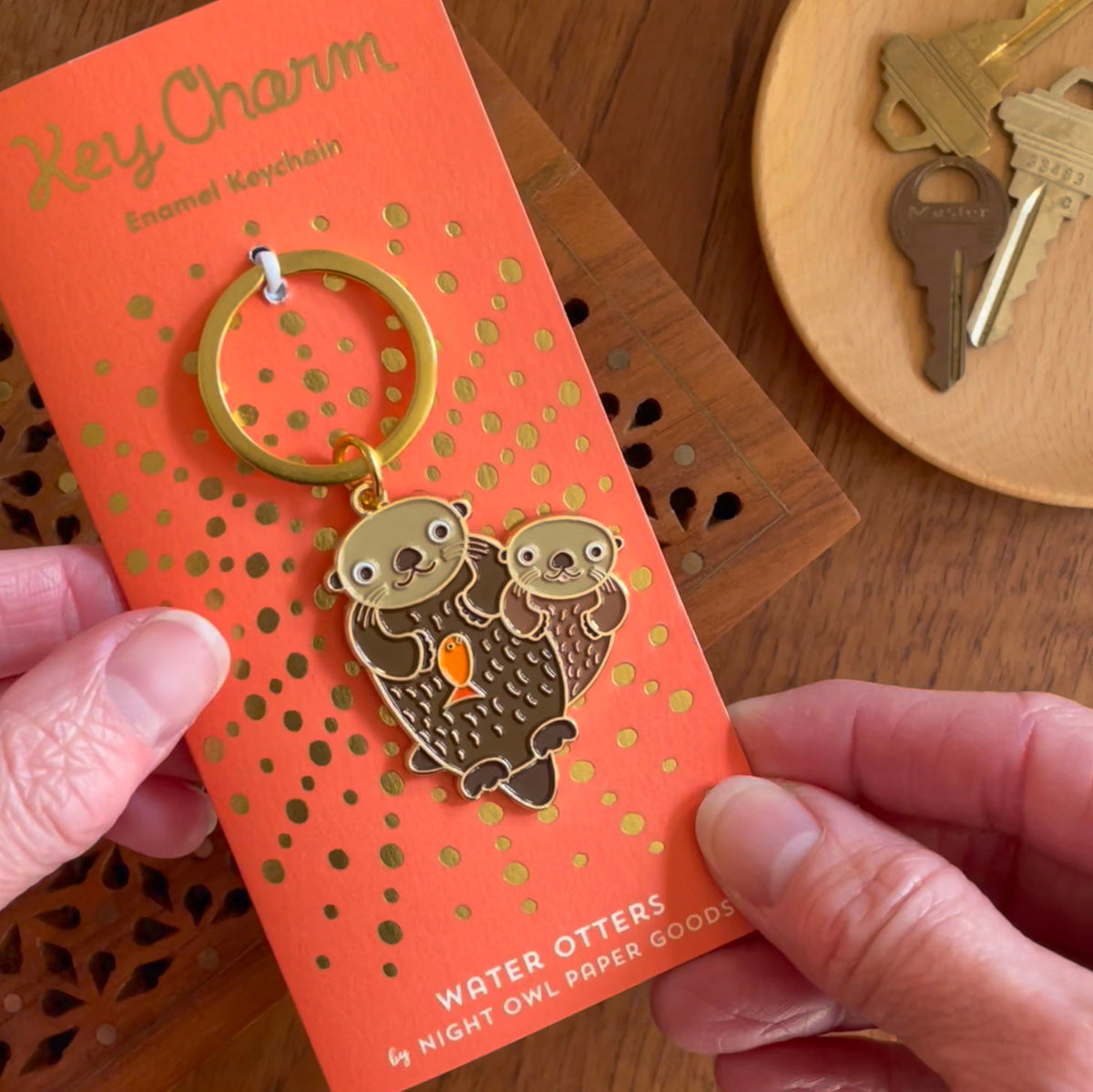 Otter enamel keychain on gold foil stamped orange backer card. Backer card reads "Key Charm Enamel Keychain" at top and "Water Otters by Night Owl Paper Goods" at bottom.