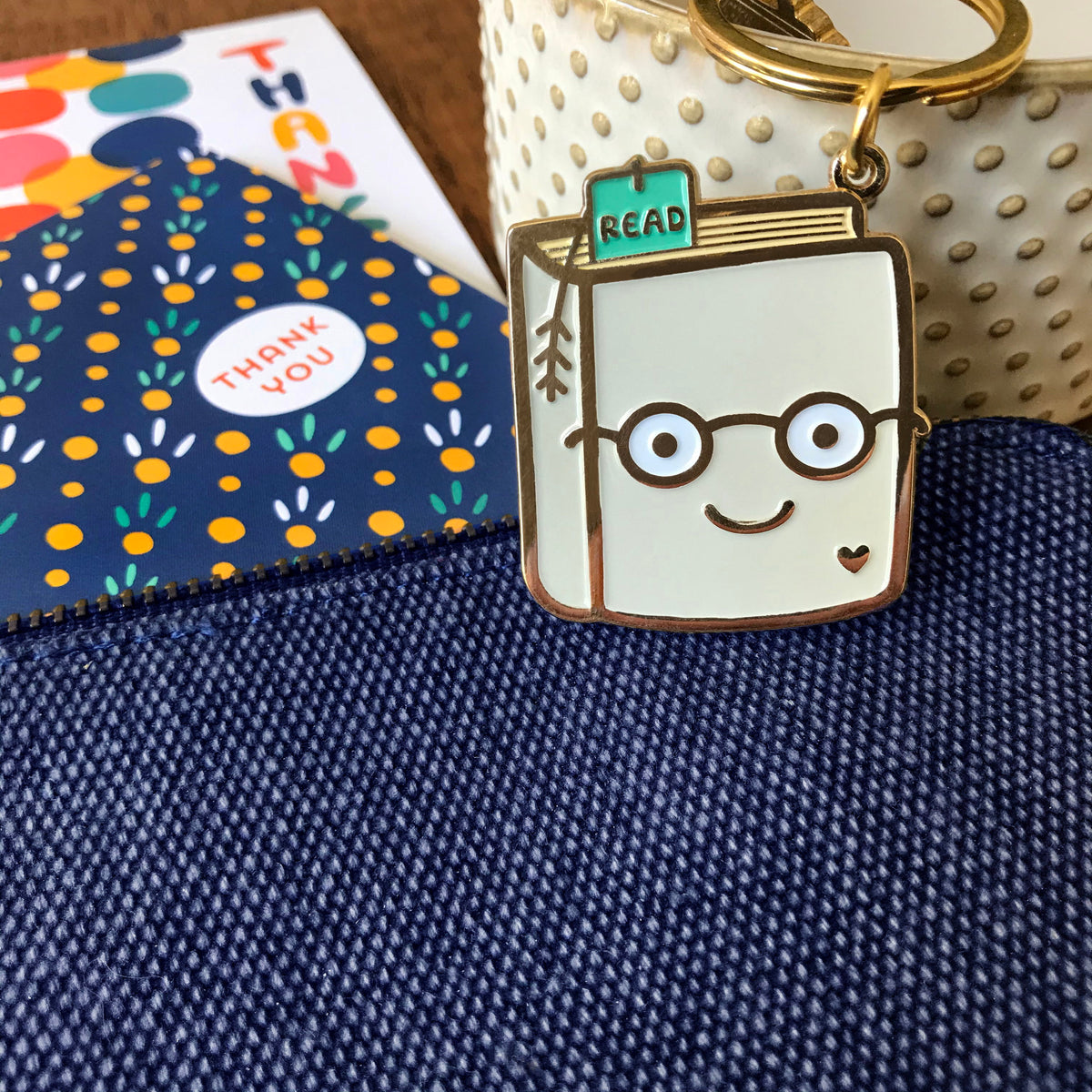 Gold metal Book Lover enamel keychain featuring a smiling book wearing round glasses with a teal "READ" bookmark and small heart detail on a gold key ring