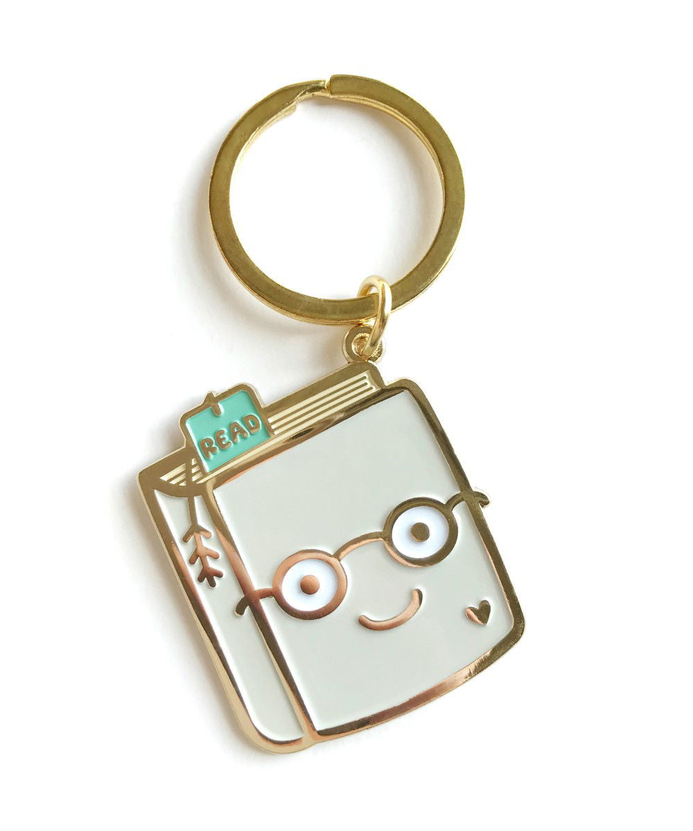 Gold metal Book Lover enamel keychain featuring a smiling book wearing round glasses with a teal "READ" bookmark and small heart detail on a gold key ring