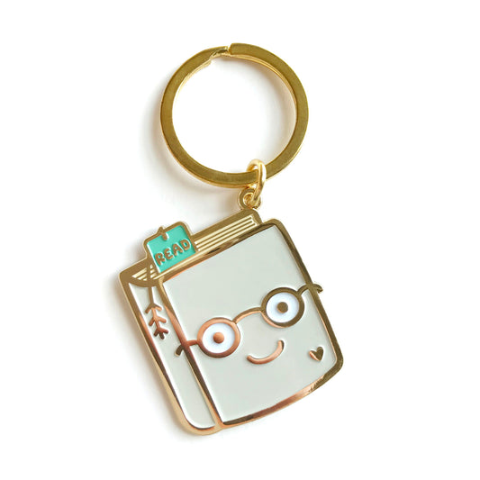 Gold metal Book Lover enamel keychain featuring a smiling book wearing round glasses with a teal "READ" bookmark and small heart detail on a gold key ring