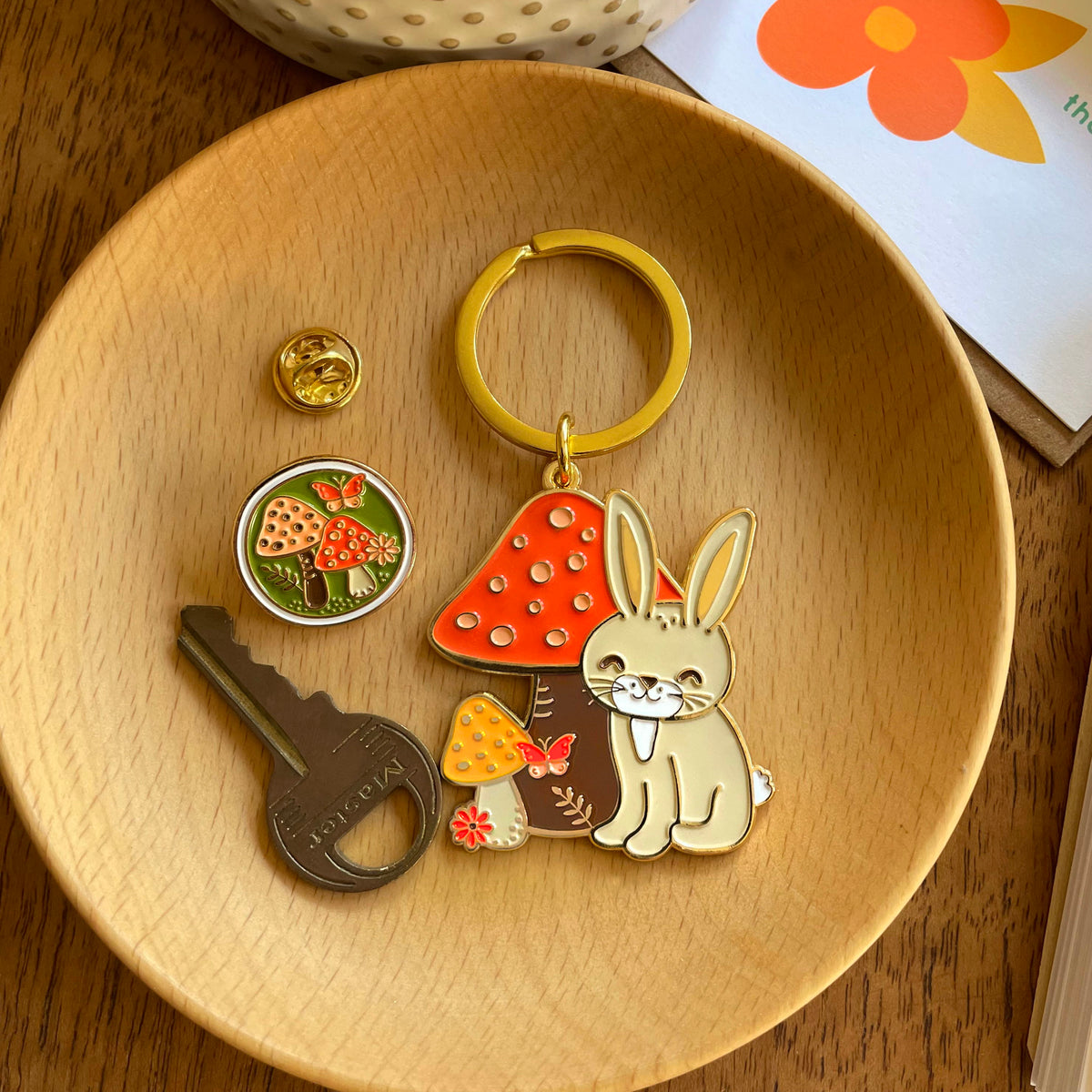 Bunny Rabbit enamel keychain in a wood bowl alongside a Mushrooms enamel pin and a gold key.