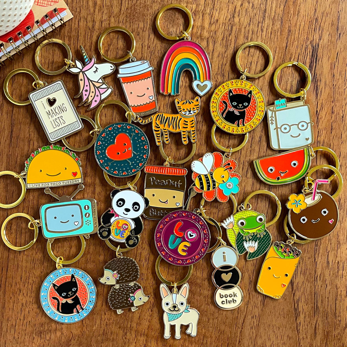 Assortment of keychains on a wood surface, some of which include a rainbow, coffee cup, unicorn, happy heart, taco, love panda, bumble bee, watermelon, coconut, frog, hedgehogs and french bulldog.