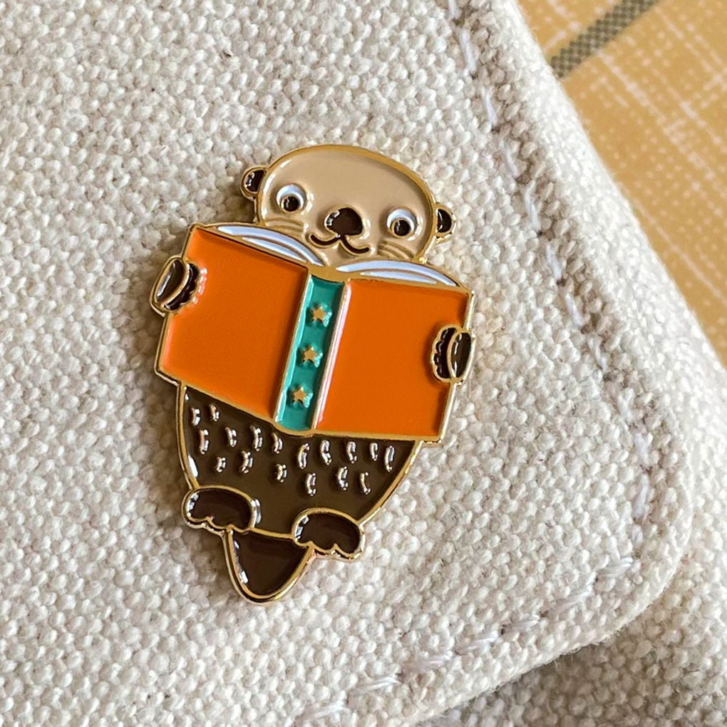 Enamel pin of an otter reading a book