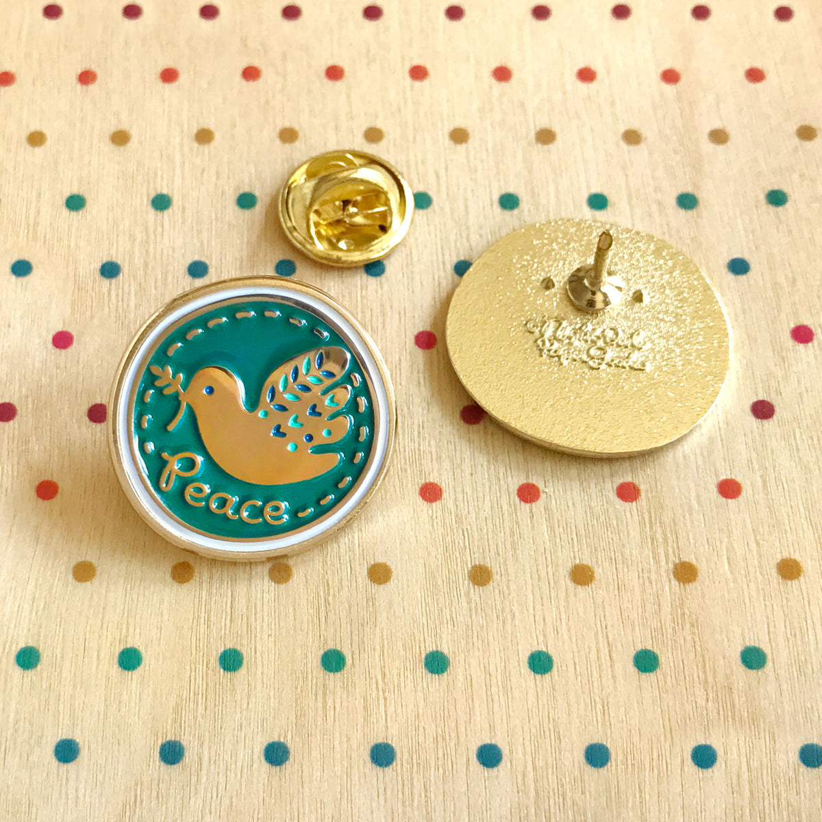 Front and back of circular shape Peace Dove enamel pin, alongside a gold butterfly clasp. Front is a teal enamel pin with a folksy gold dove holding an olive branch in its beak and gold 'Peace' text. Back is gold with Night Owl Paper Goods script logo. 