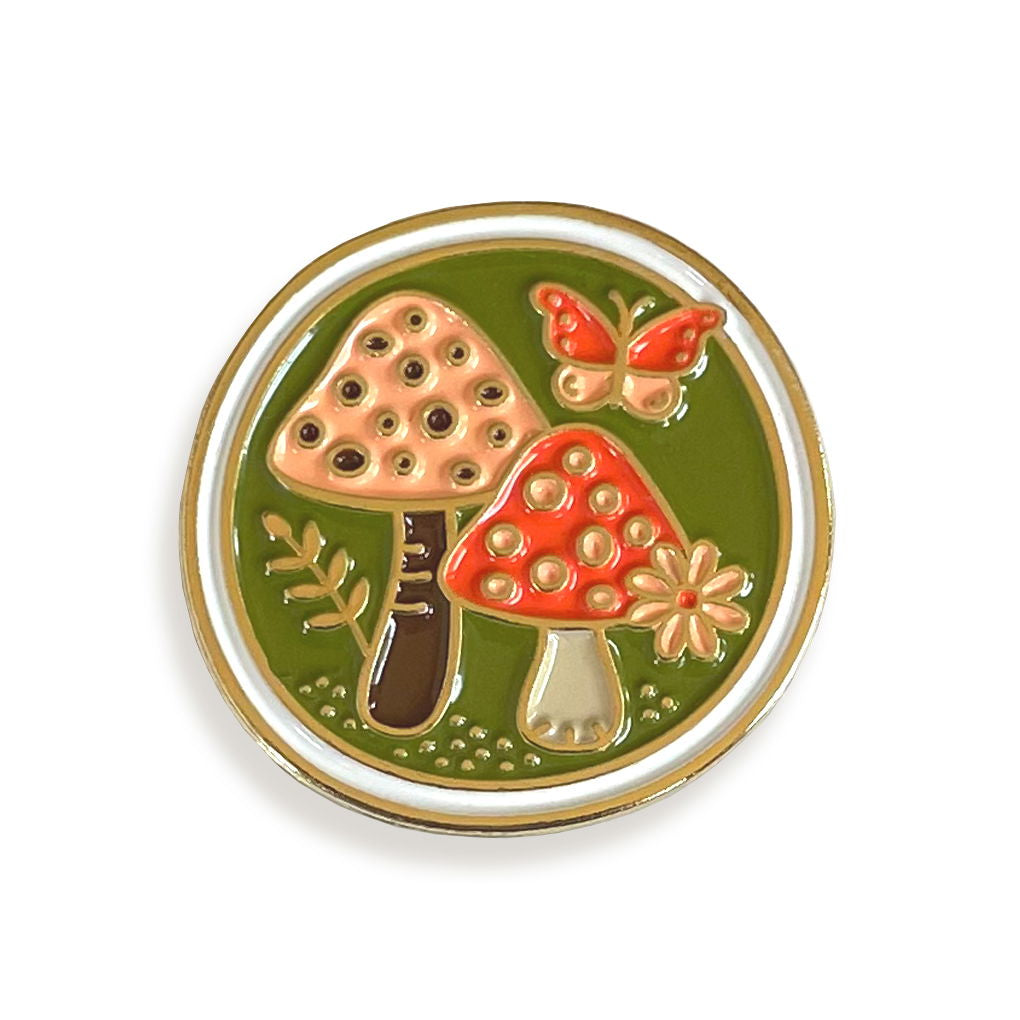 Round enamel pin with red and peach mushrooms, a butterfly and a flower on a green background, bordered with gold details.