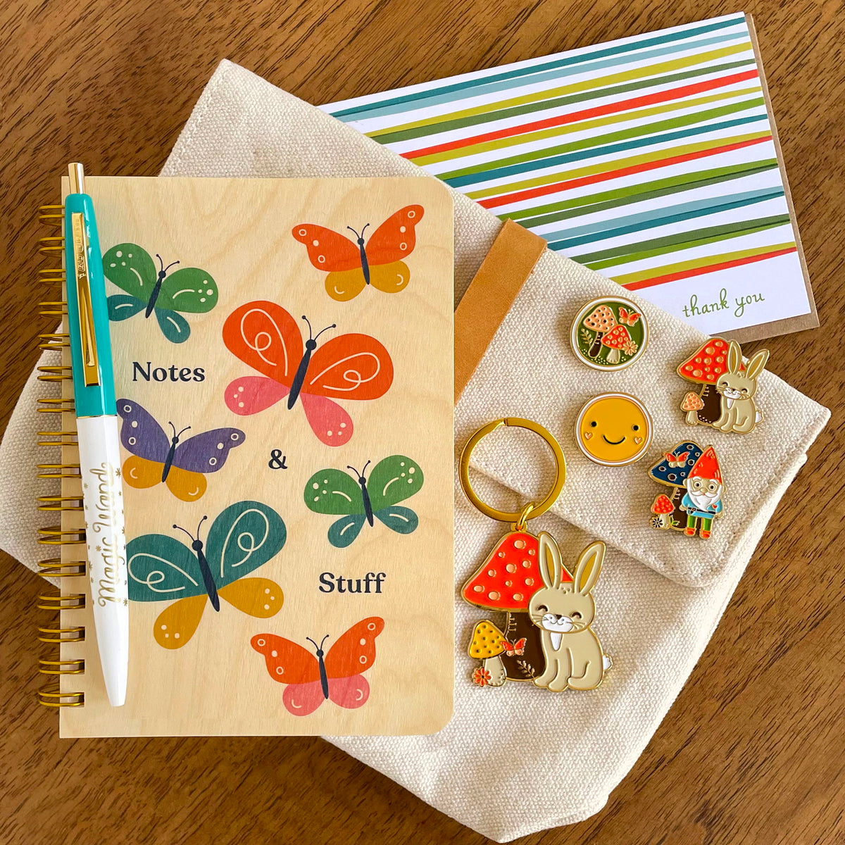 Butterfly wood notebook, Magic Wand pen, Lively Stripes Thank You card and Rabbit keychain alongside Mushroom, Rabbit, Smiley Face and Gnome enamel pins. 