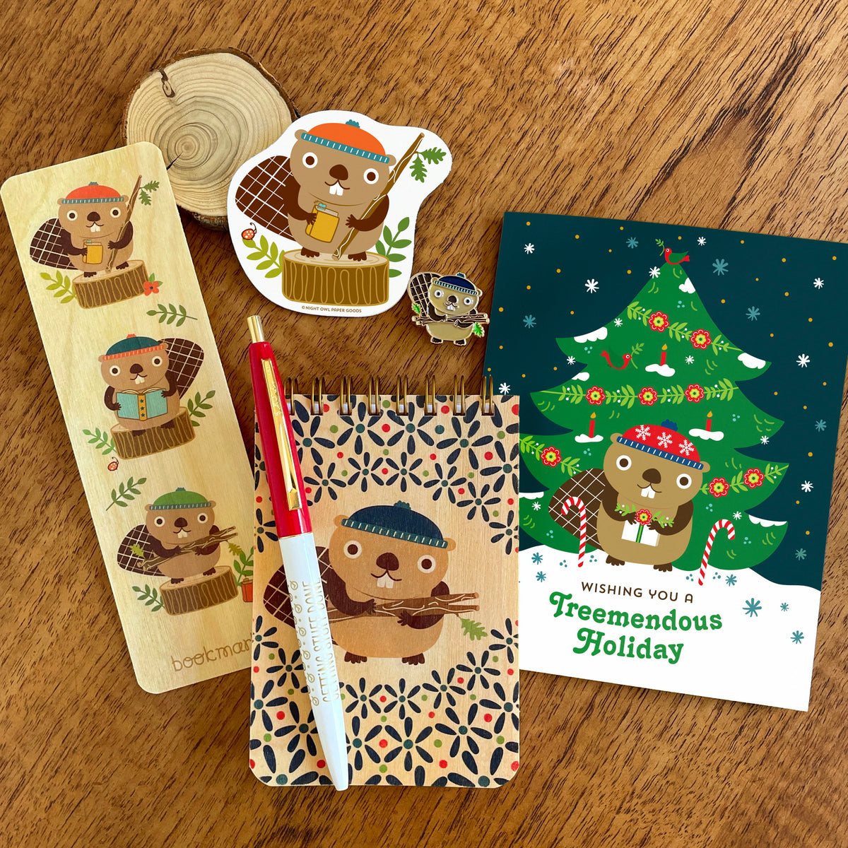 Collection of beaver items, including a wood bookmark, wood mini notepad, sticker, enamel pin and holiday card along with a red and white Getting Stuff Done ballpoint pen.